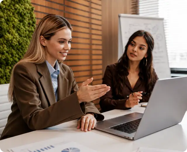 Two businesswomen reviewing financial data on a laptop screen during a collaborative office meeting.