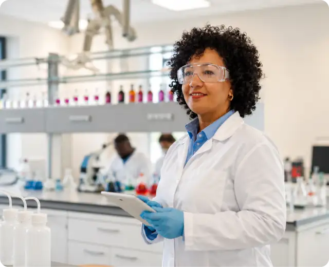 A scientist wearing a white lab coat, safety glasses, and blue gloves holds a digital tablet inside a laboratory with flasks and equipment in the background.