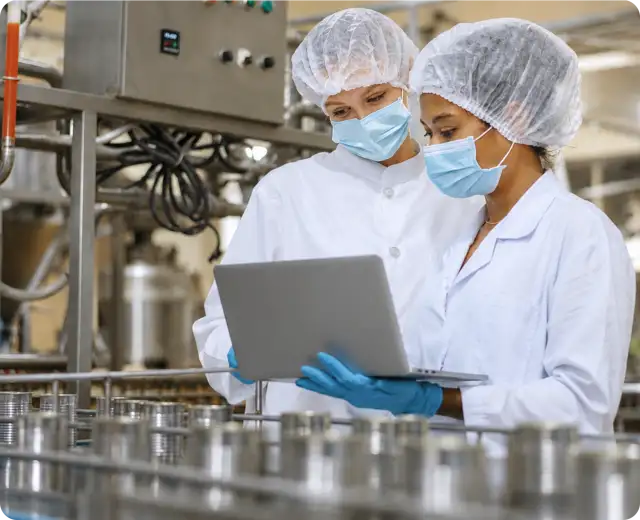 Two facility workers wearing hairnets, protective face masks, and blue gloves look at a laptop screen next to a canned food production line.