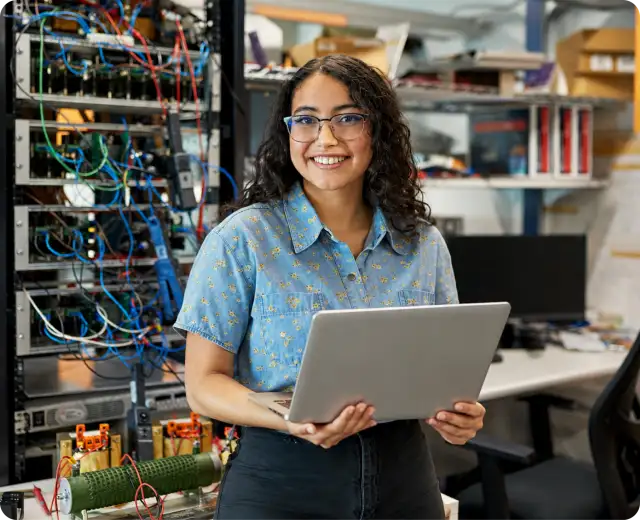 A smiling woman with glasses holds a laptop while standing in front of a server rack filled with colorful wires and electronics equipment.